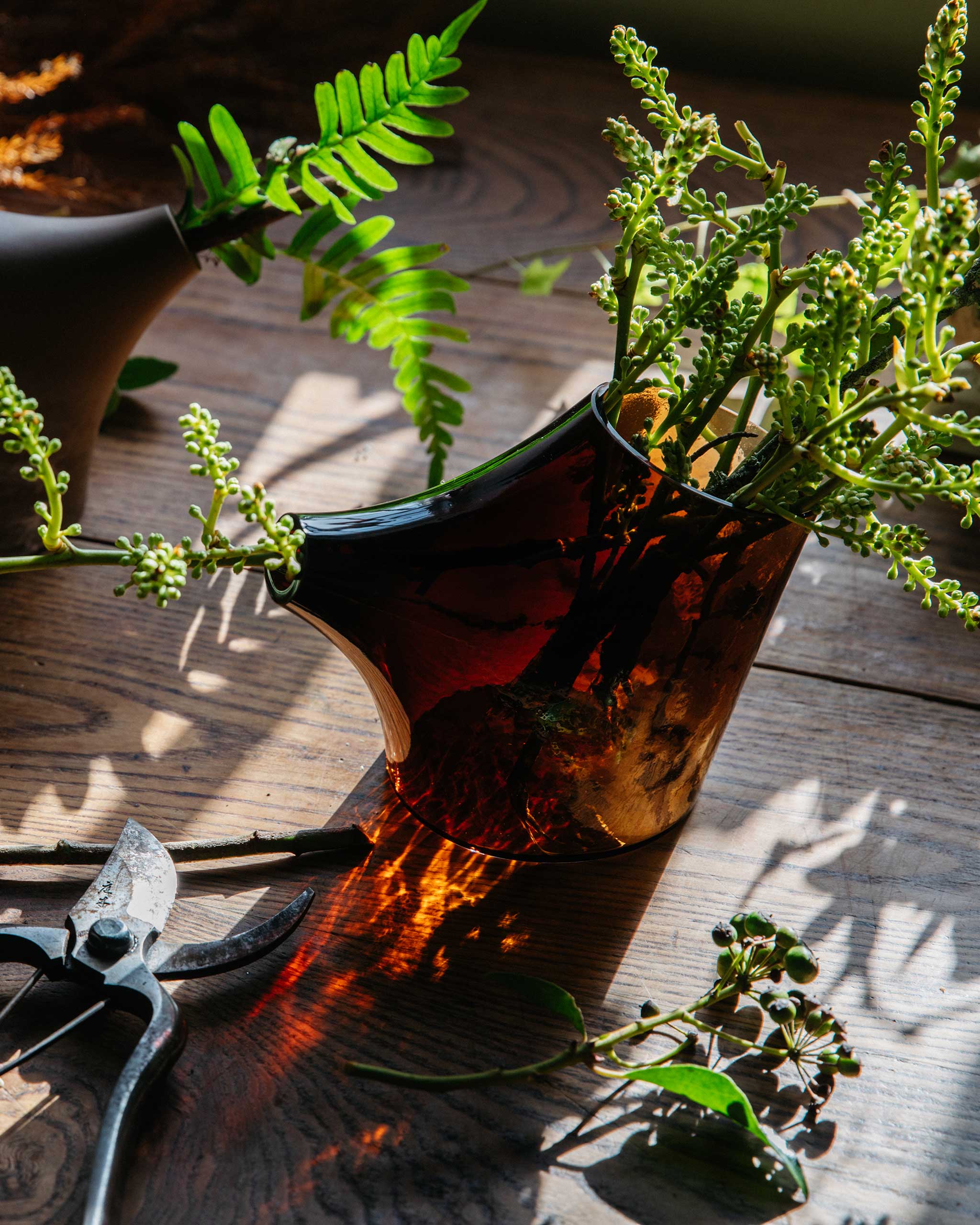 Kiln-cast blown glass pot on a warmly lit table, with light filtering through the translucent form and foliage inside, showcasing craftsmanship and material depth.