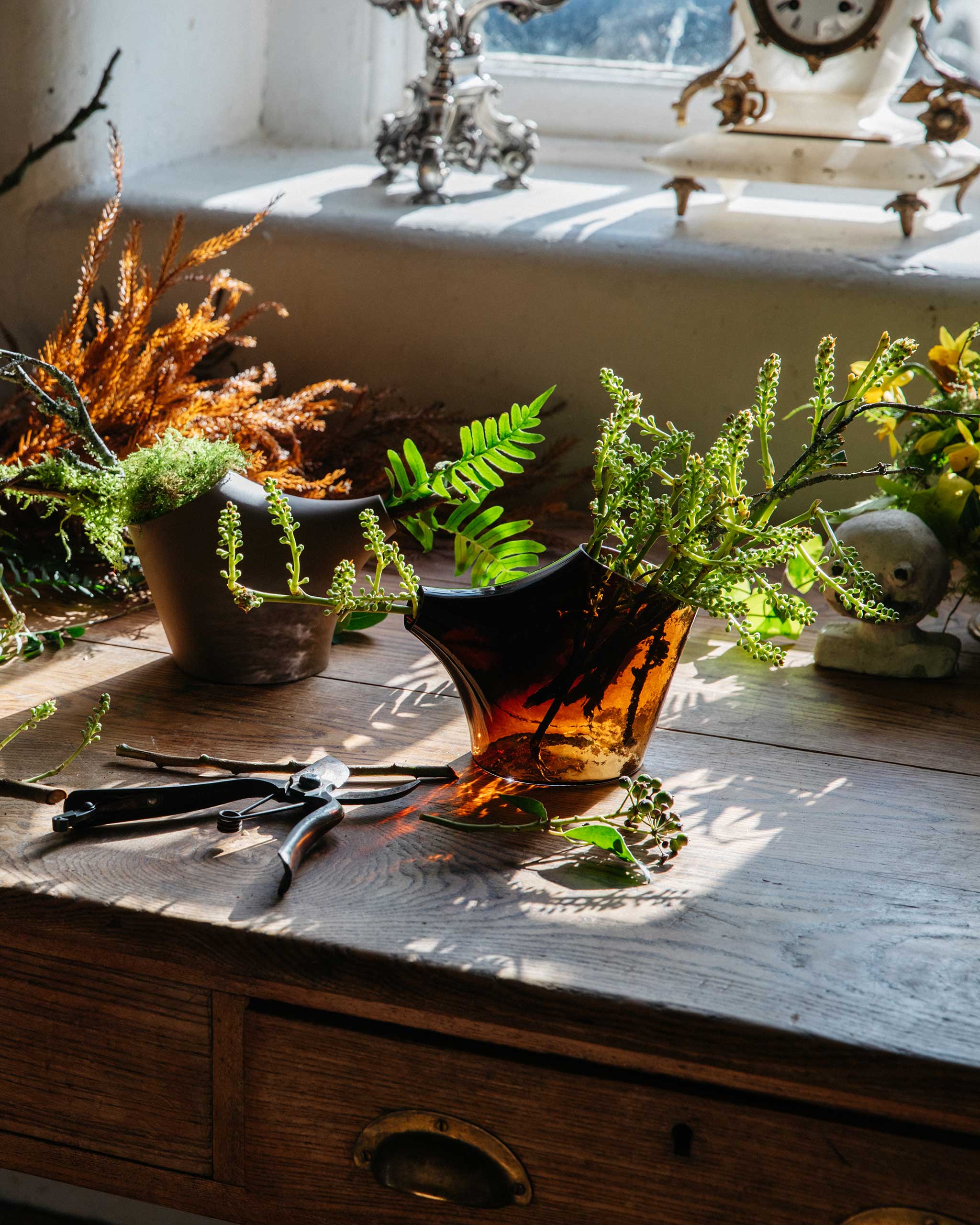 Kiln-cast blown glass pot and brown porcelain pot on a warmly lit table, with light filtering through the translucent form and foliage inside.