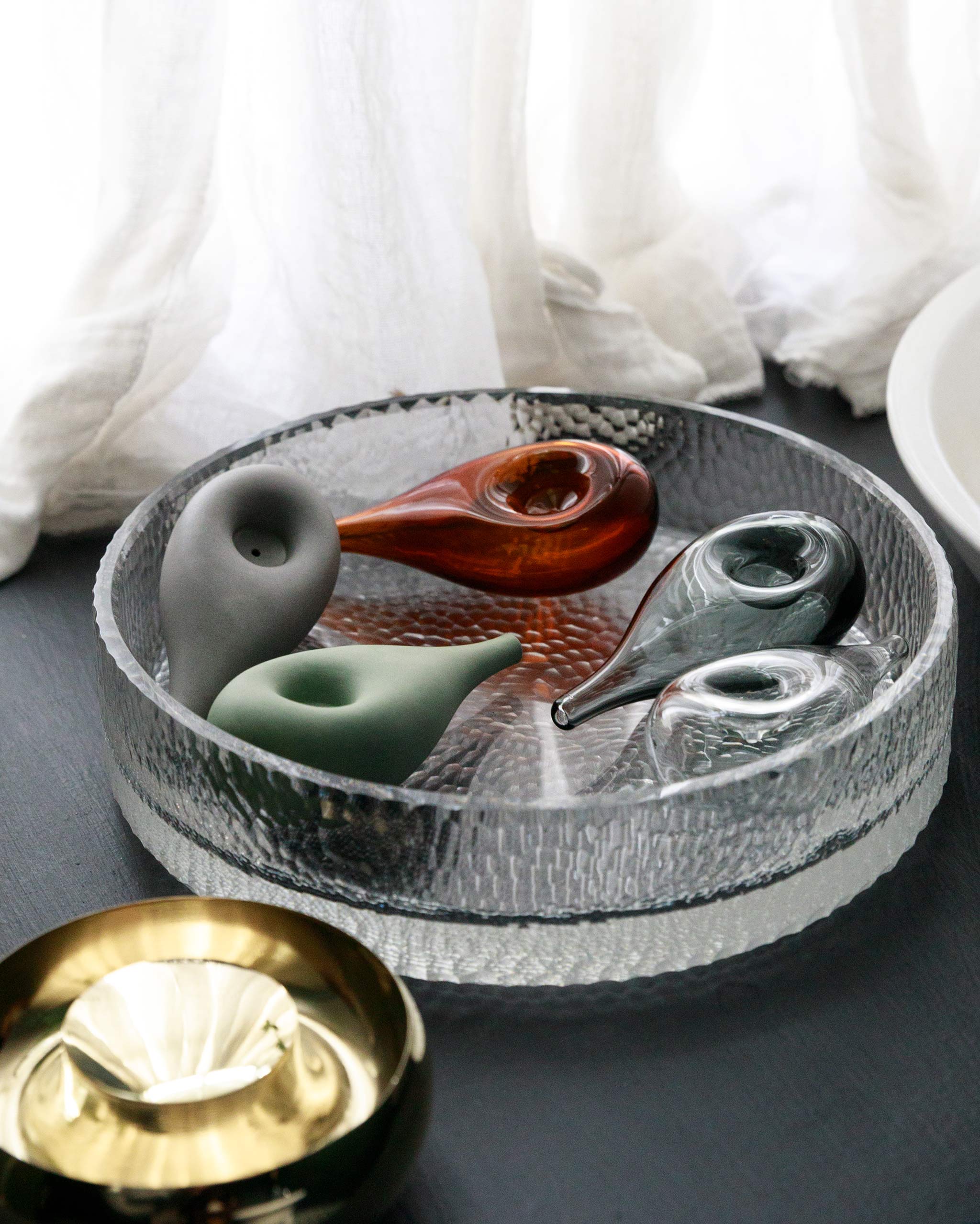 Five variations of Cloud Pipe in glass and porcelain arranged in a crystal bowl with a gold vessel in front, softly lit against white curtains.