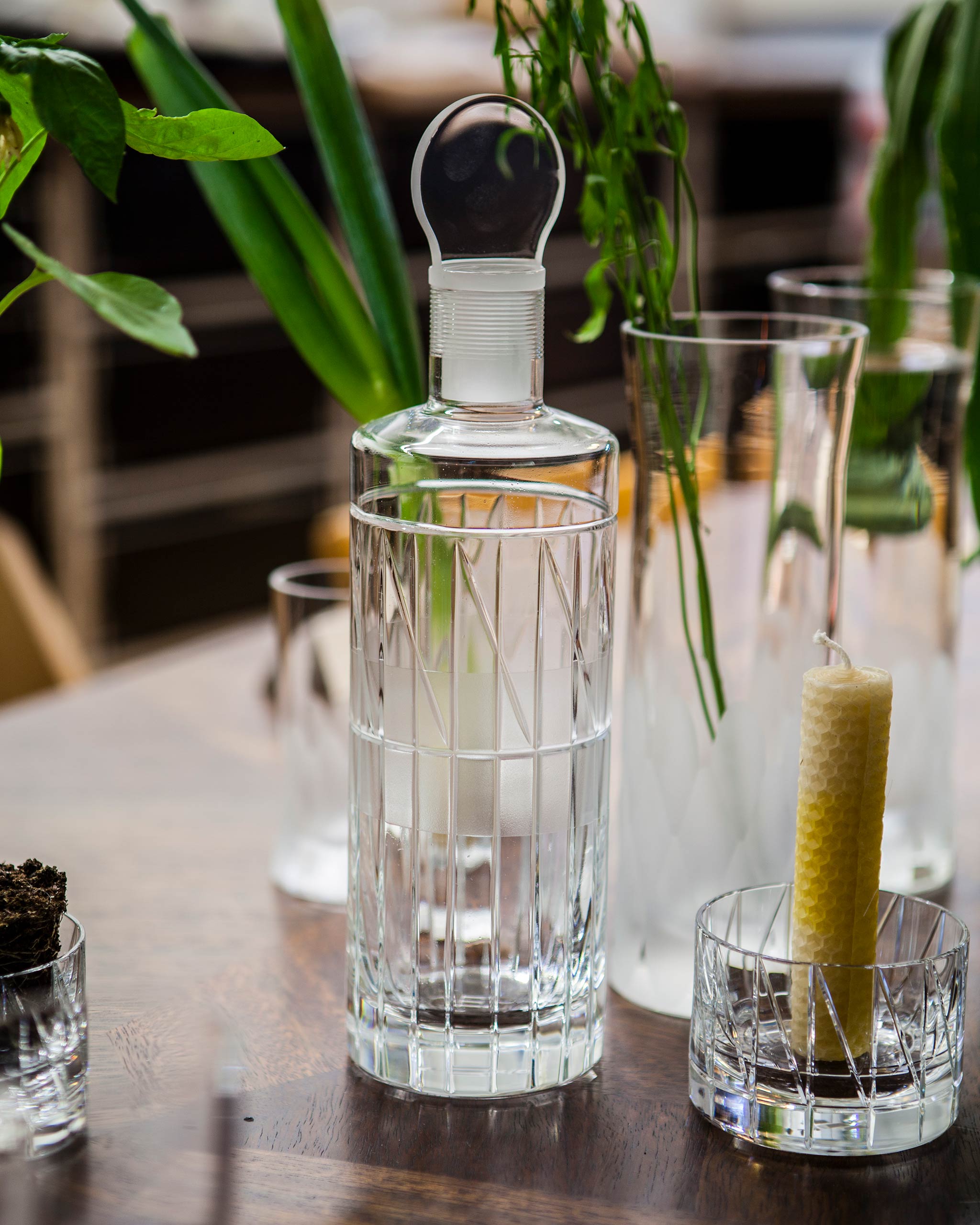 Hand-blown whiskey decanter with stopper, modern cut crystal design sitting on a table alongside other crystal glassware and foliage.
