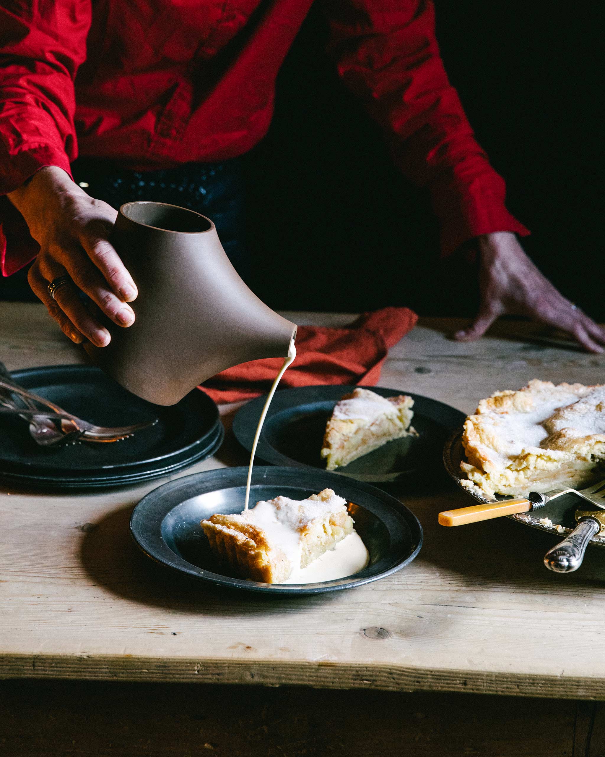 Lifestyle image of a brown porcelain pot holding custard, the custard being poured over a piece of pie.