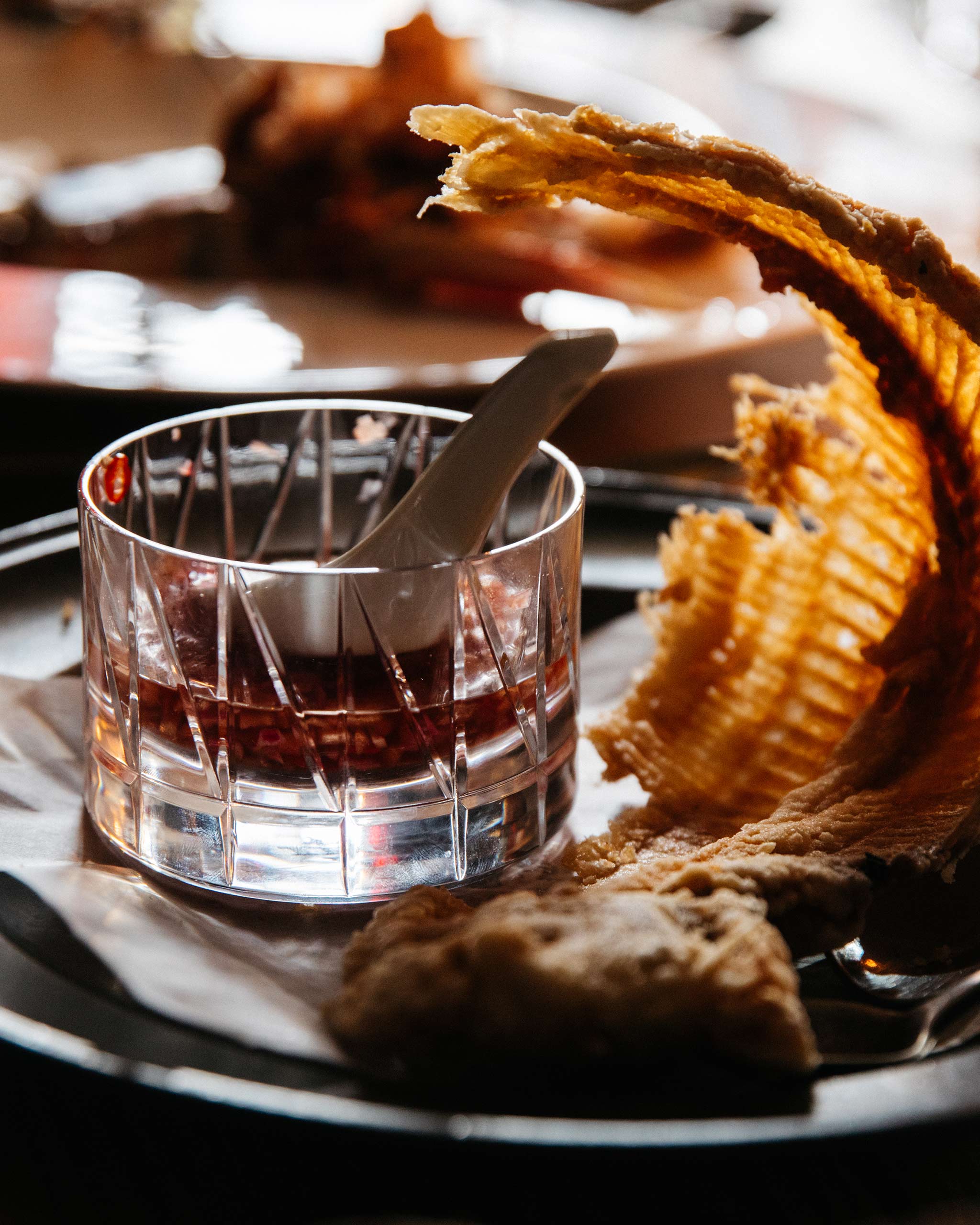 A dining scene with a hand-cut crystal low whiskey glass holding a sauce and serving spoon.