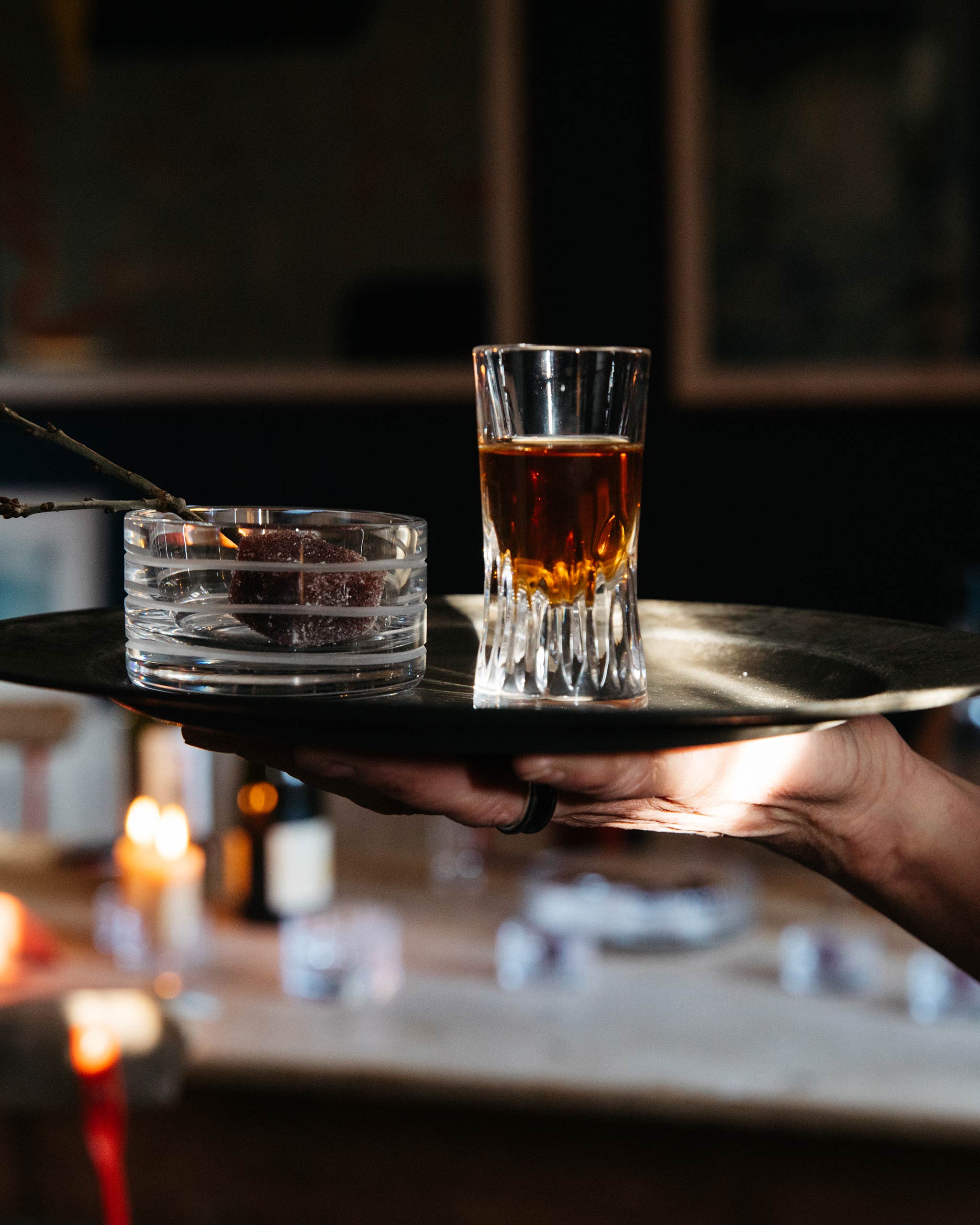 Lifestyle image of a held plate on which stands a hand-cut crystal shot glass containing whiskey alongside a votive holding a sweet.