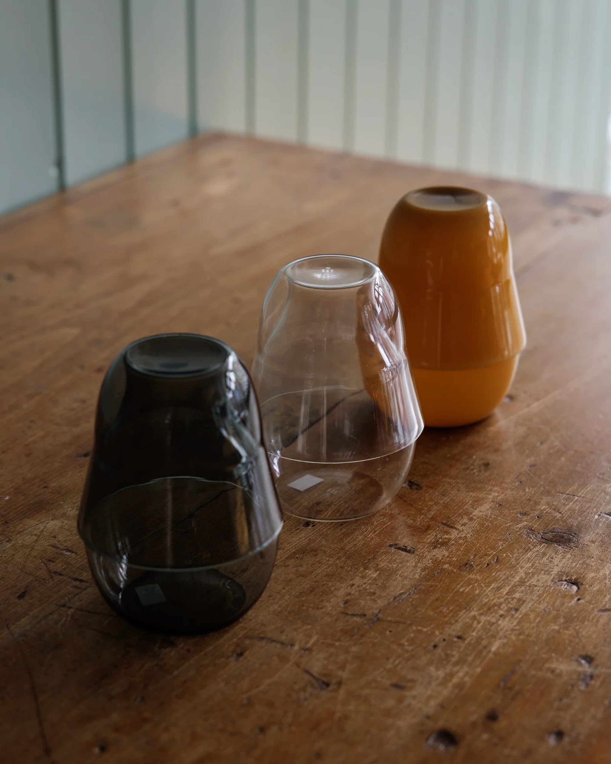 Set of three mouth-blown borosilicate glass carafes with glasses stacked on top, in ochre, grey, and clear finishes, displayed on a wooden table in natural light.