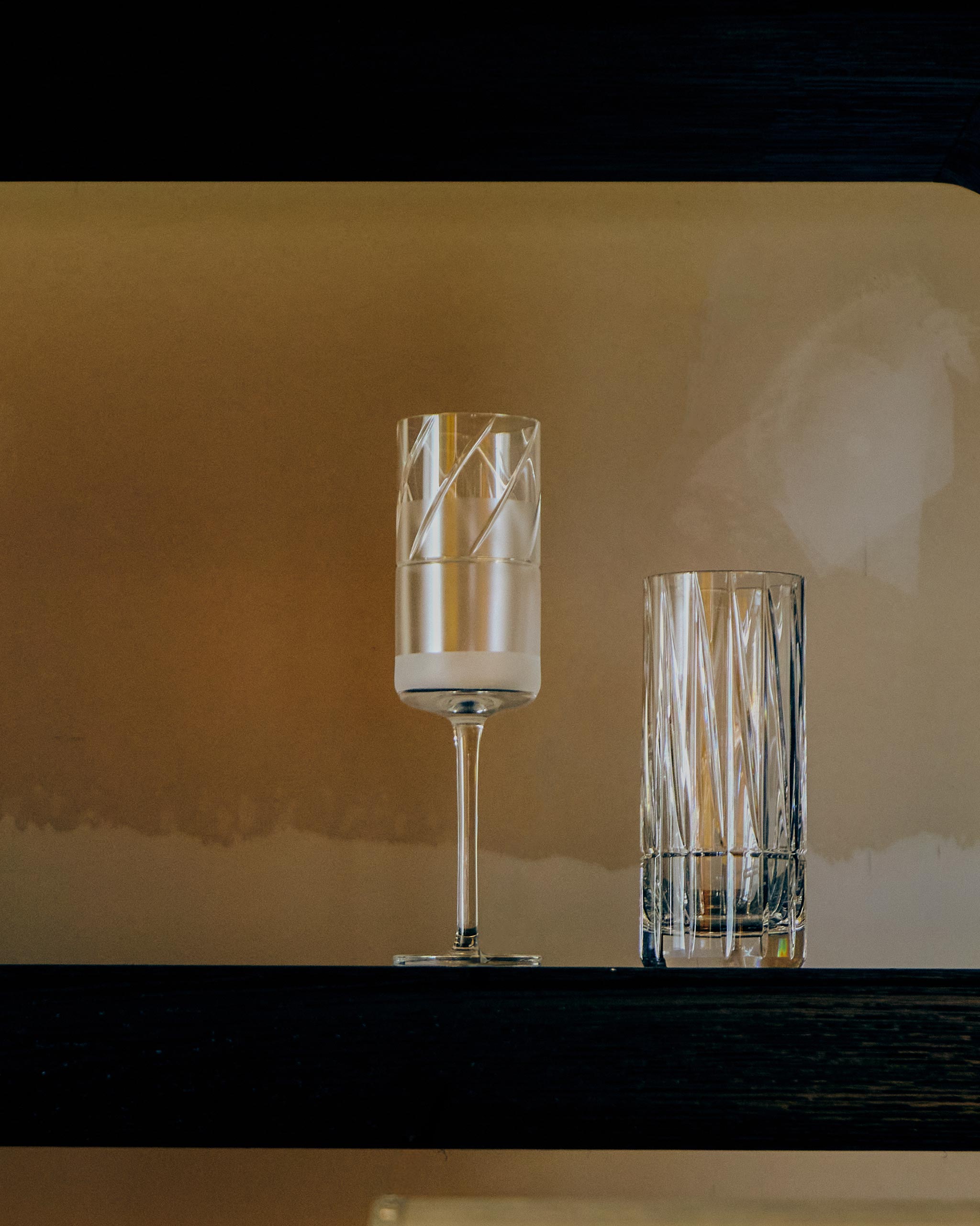 Crystal champagne flute displayed beside highball glass on a wooden shelf, softly lit to highlight sandblasted detailing.