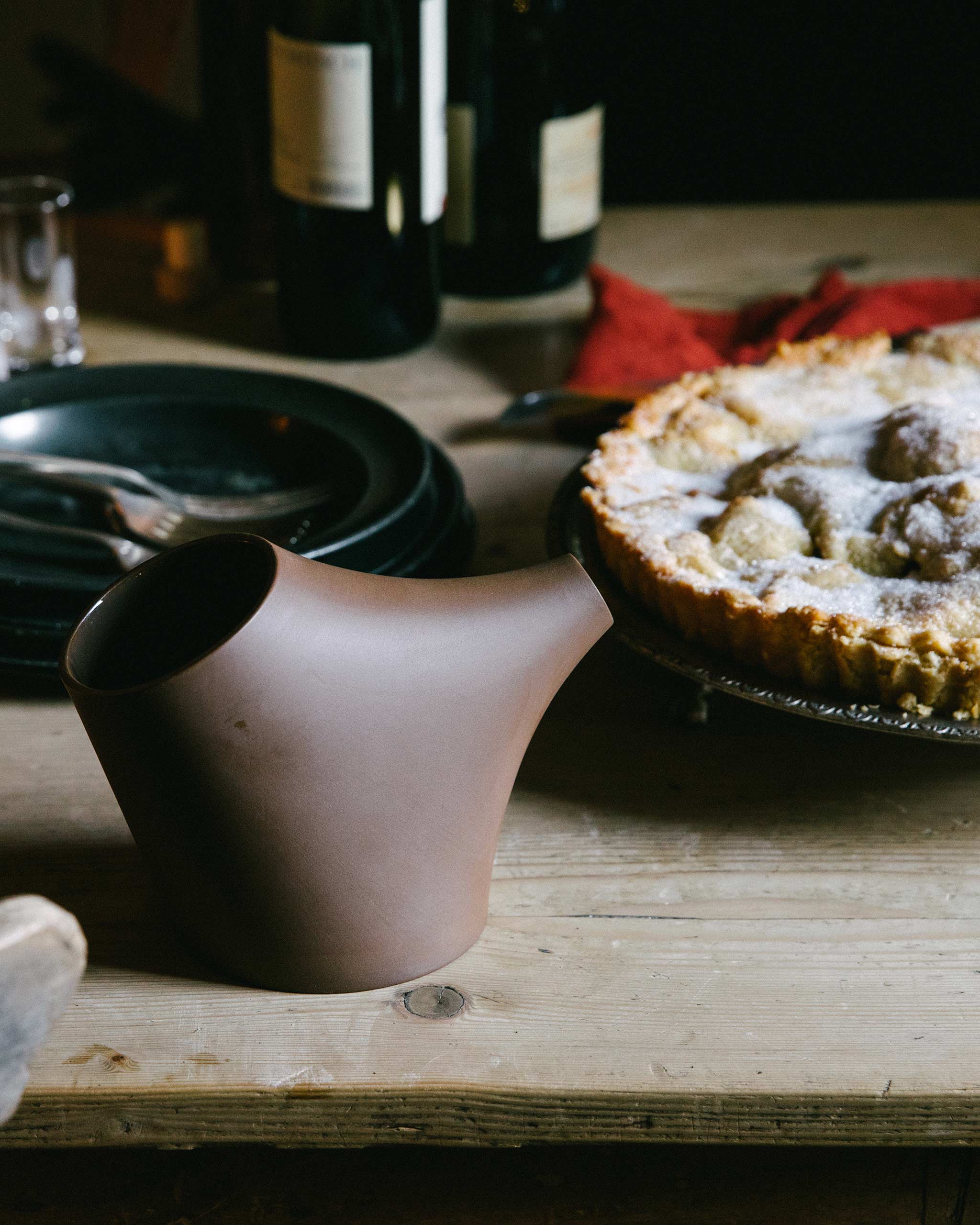 Lifestyle image of a festive table on which there is a brown porcelain pot, a pie, wine glasses and cutlery.