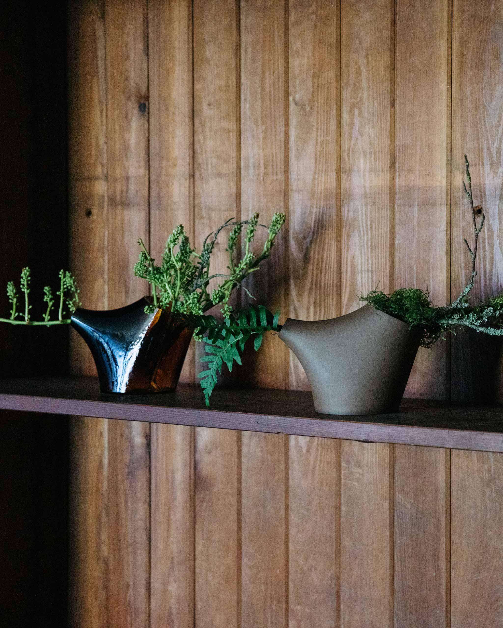Two HopStep pots displayed on a wooden shelf, each holding natural foliage, highlighting variations in form, tone, and craftsmanship across the collection.