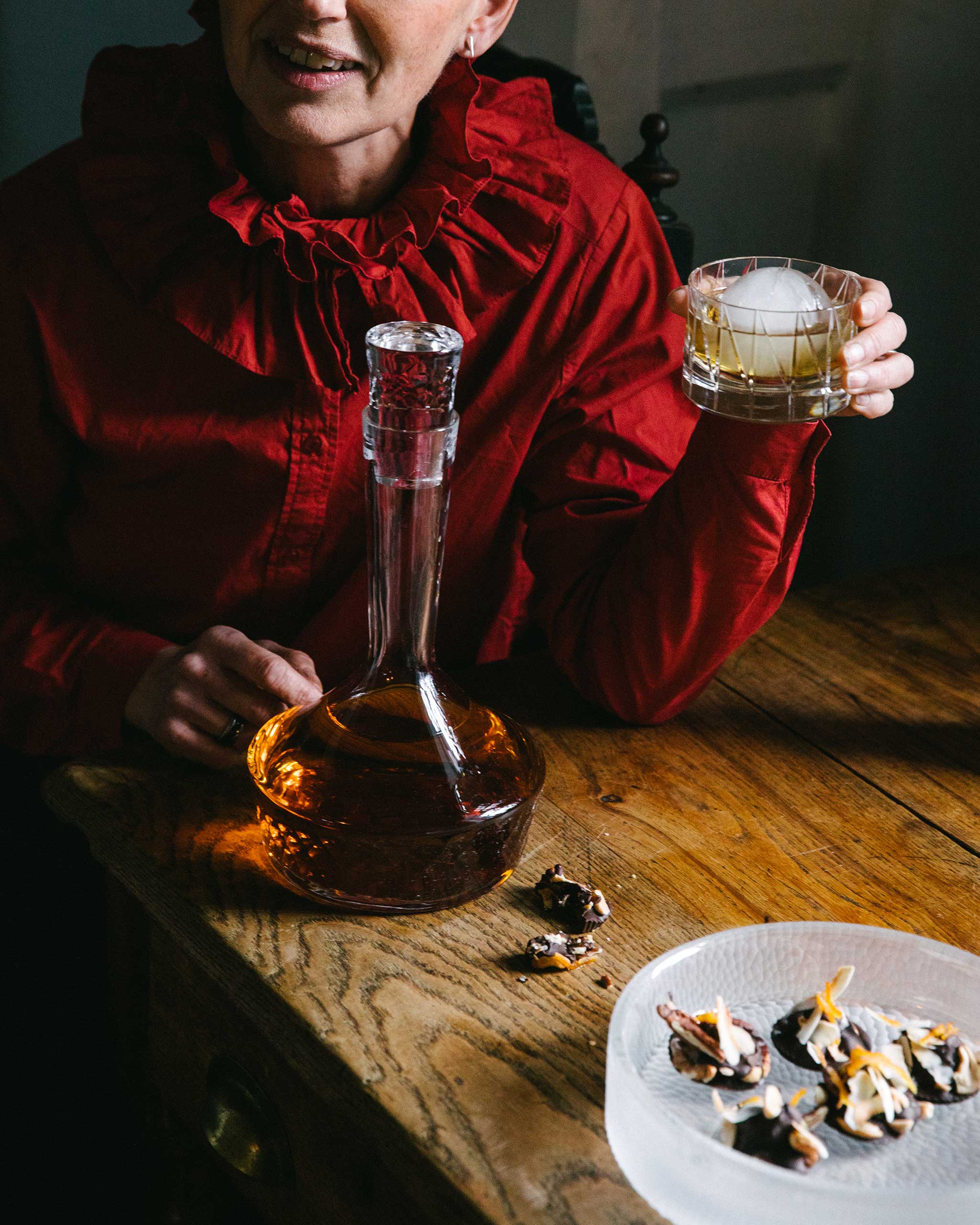 Cut crystal low whiskey glass with large ice-cube held in a woman's hand. A hand-cut crystal decanter sits on the table.