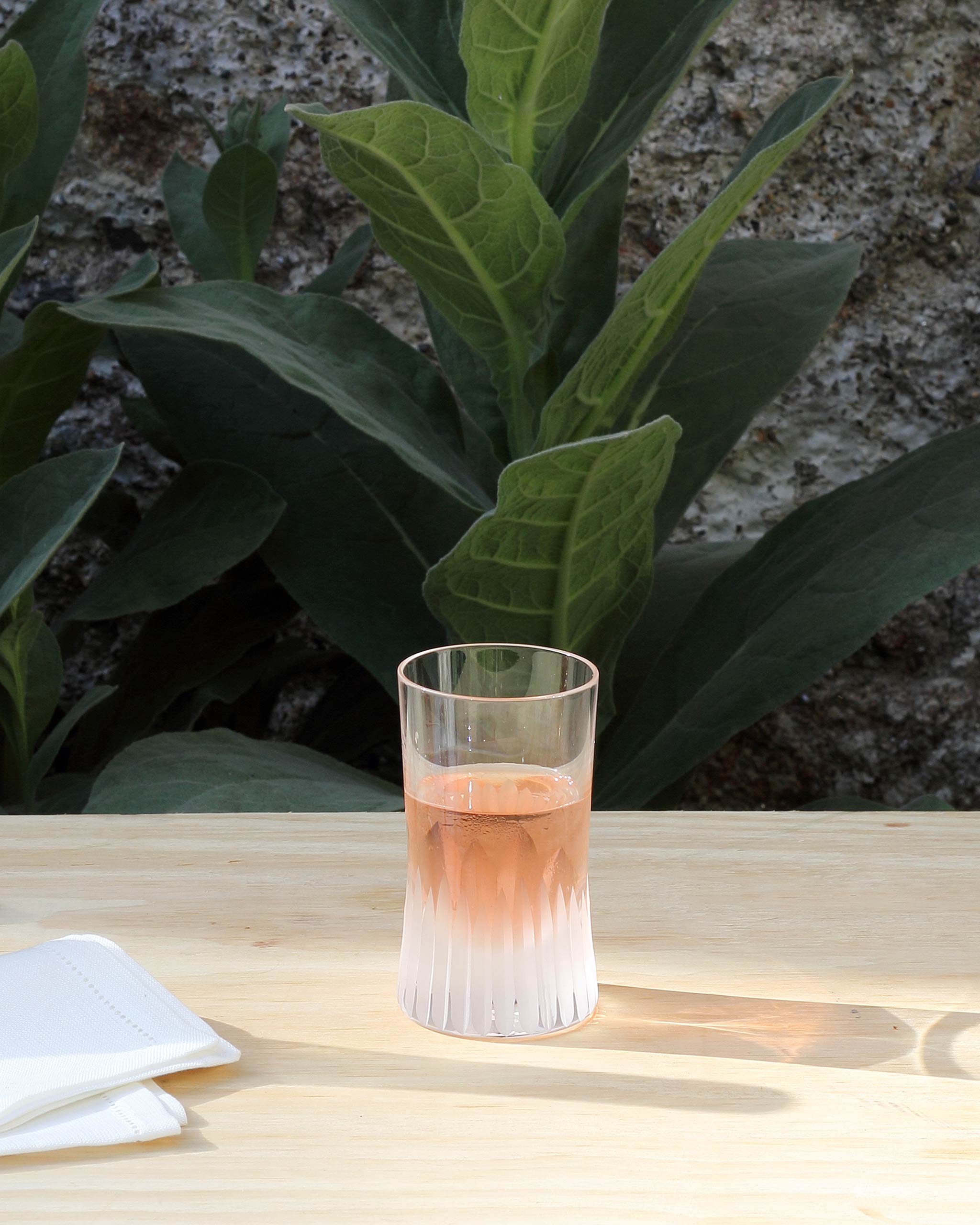 Lifestyle imagery of an unpolished hand-cut Irish crystal rocks tumbler glass with deep angular cuts sitting on a table in front of natural foliage