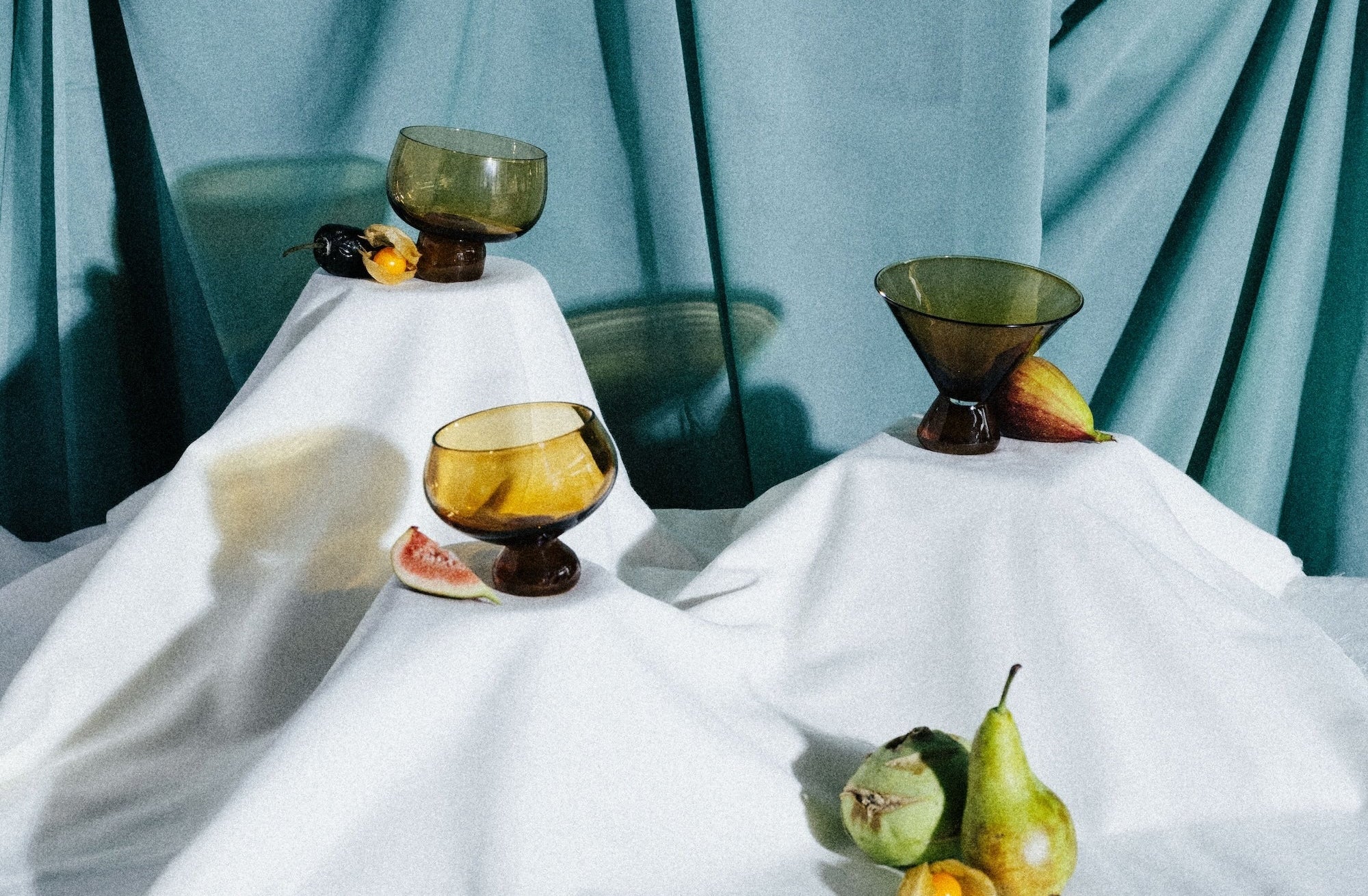 Three vintage-style glass goblets on a white tablecloth with fruits against a teal curtain background.